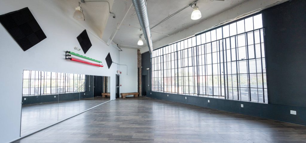 A spacious dance studio with a large mirror covering one wall, barres along another, and a high ceiling with exposed ductwork. The room is lit by natural light coming through the tall windows that cover the entire back wall.