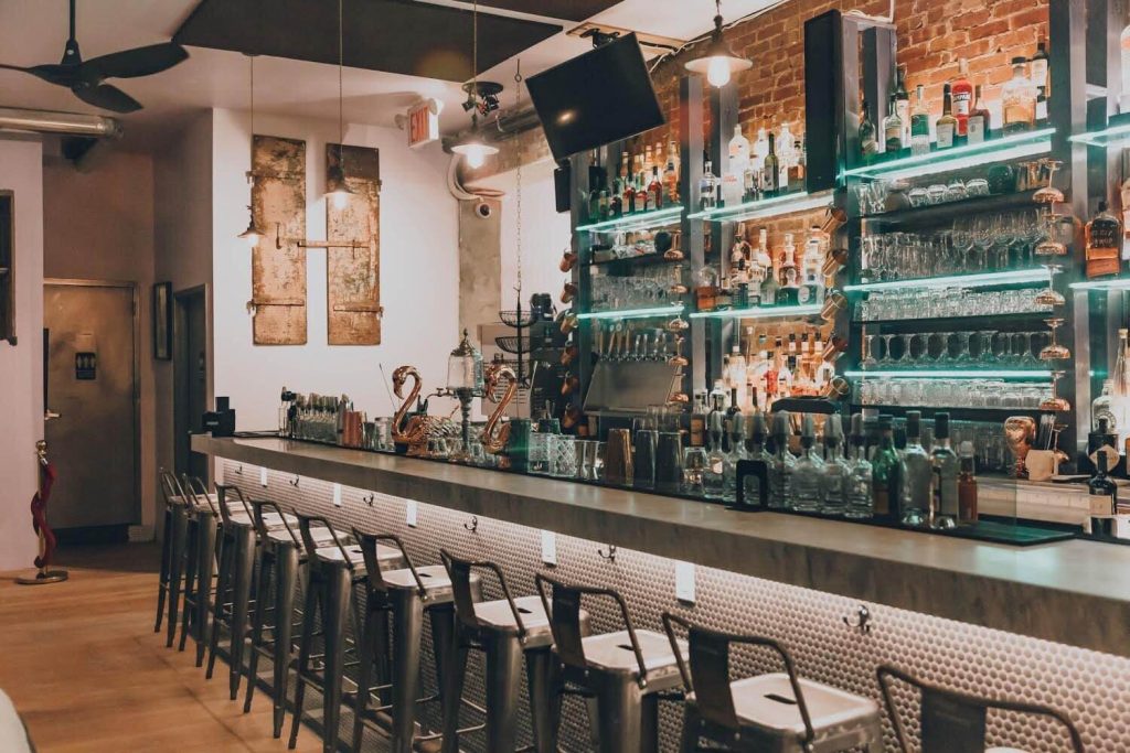 A modern bar with exposed brick and lit glass shelves full of various bottles, barstools lined up along the bar counter, and pendant lights hanging from the ceiling, creating a warm and inviting atmosphere.