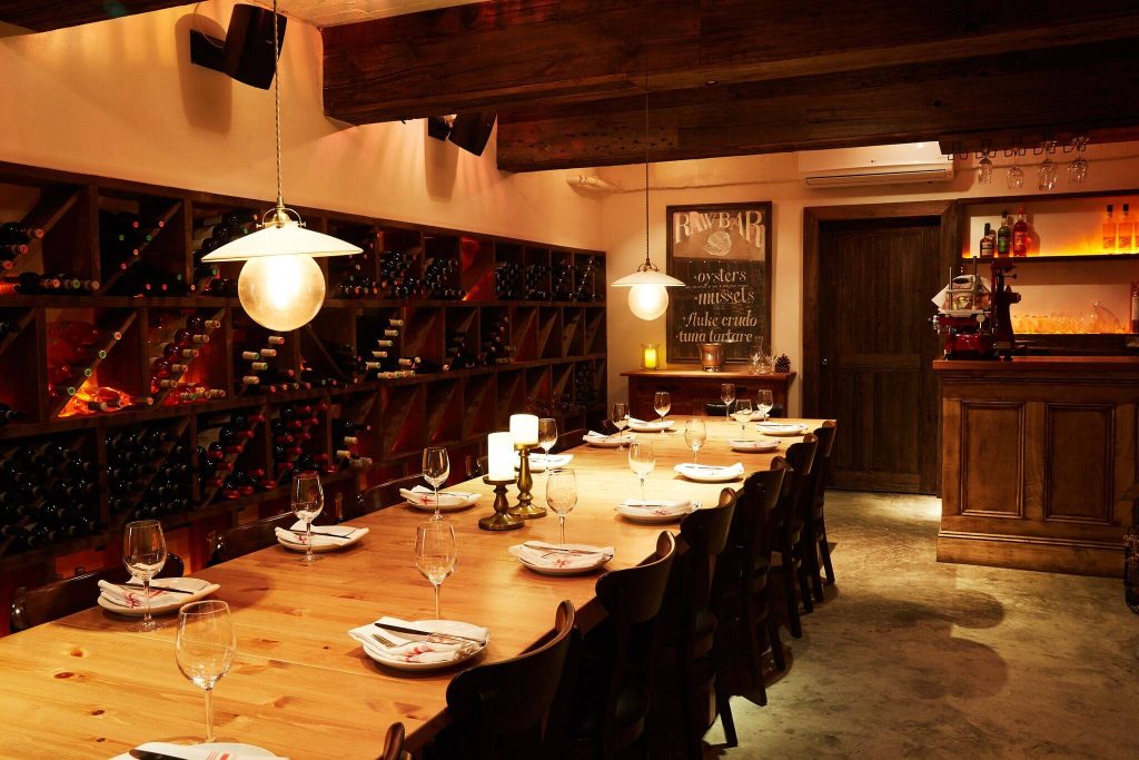 A private dining room with a long wooden table, elegant glassware, wine bottles on shelves, and soft pendant lighting, ideal for an intimate birthday dinner.
