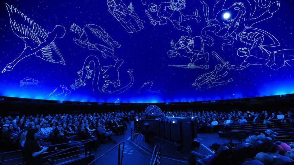 A planetarium with a darkened dome ceiling displaying illuminated constellations and a seated audience looking up, enjoying an educational and immersive experience.