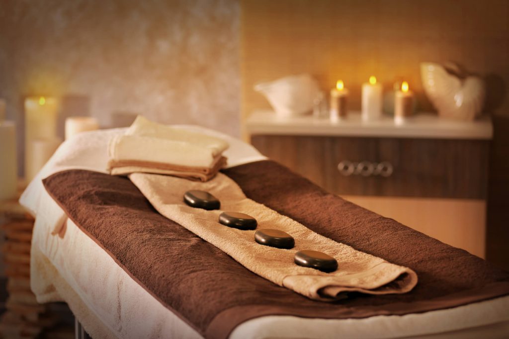 A spa treatment room with a massage table set with towels and hot stones, soft lighting, and candles, conveying a serene and relaxing ambiance for a couple's massage.