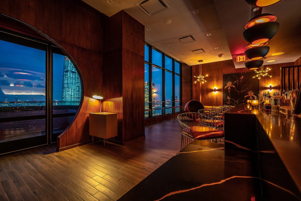 A chic bar with modern decor, dim lighting, and a panoramic window offering a night view of the city skyline, creating an upscale and romantic atmosphere.