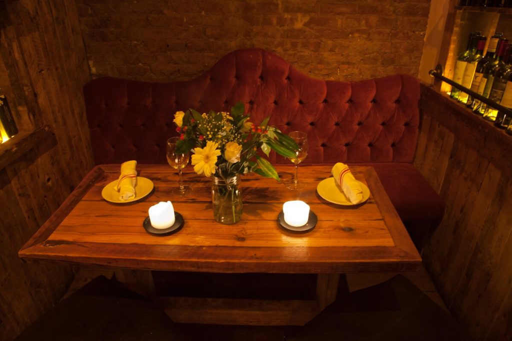 A cozy corner of an Italian restaurant with a rustic wooden table, plush red velvet seating, lit candles, and a bouquet of flowers, suggesting an intimate dining experience.