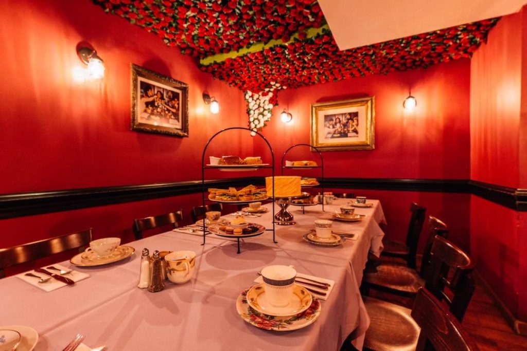 An elegantly set table for a high tea party in a room with red walls and ceiling decorations of green and red flowers. Framed pictures hang on the walls and the table is laid with fine china and an array of tea snacks.