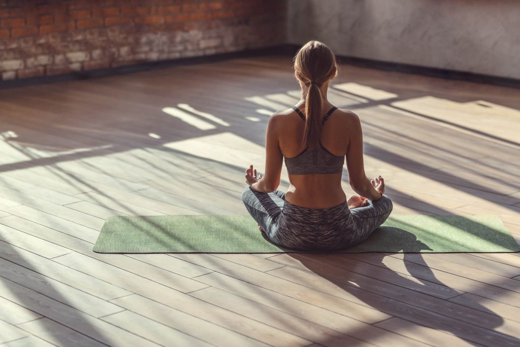 
A woman is seated on a yoga mat in a cross-legged meditation pose, with her back to the camera. She is indoors, in a room with wooden flooring and a brick wall, lit by natural sunlight streaming through windows, casting shadows on the floor. She is wearing a gray sports bra and patterned leggings, with her hair tied up neatly, indicating a tranquil and focused yoga session.