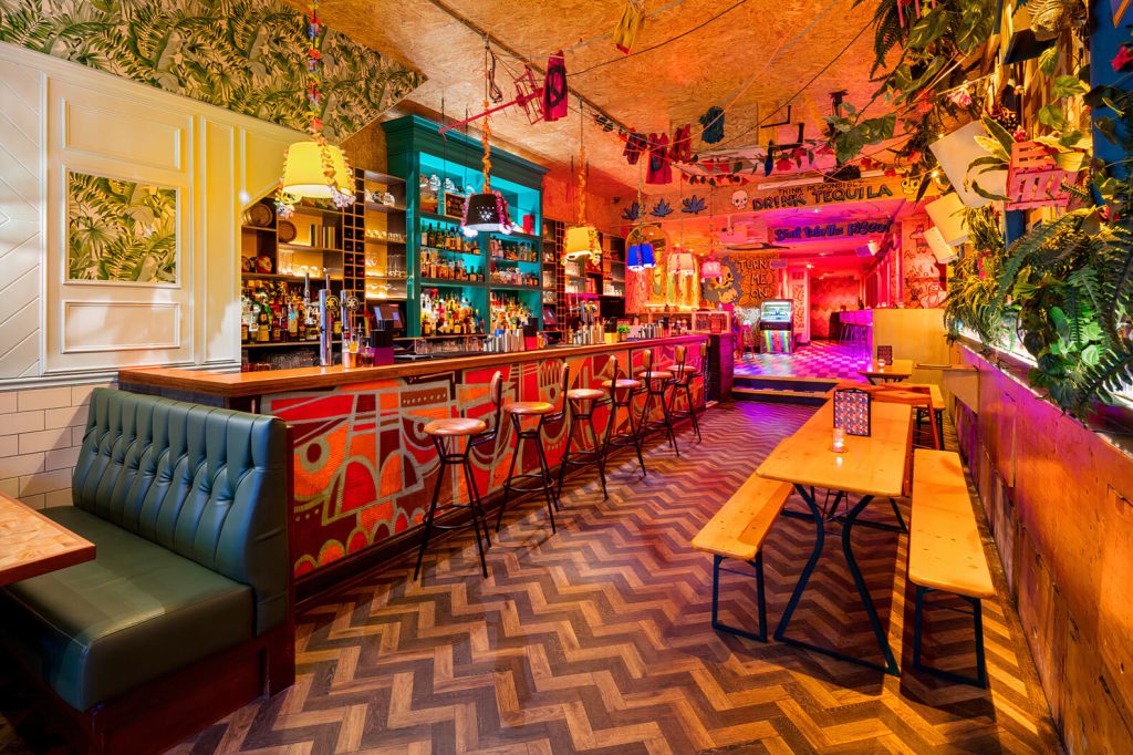 A lively, colourful bar with eclectic decor, festooned with bright lights and playful artwork, creating a festive and fun atmosphere.