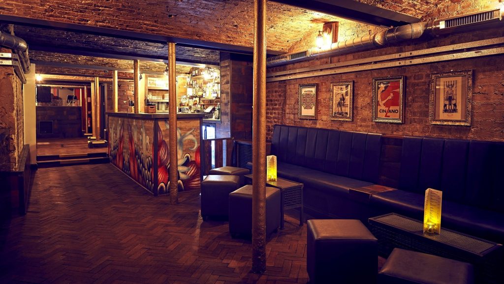 A rustic bar with exposed brickwork, herringbone wooden floors, and atmospheric lighting, perfect for a cosy evening with friends.