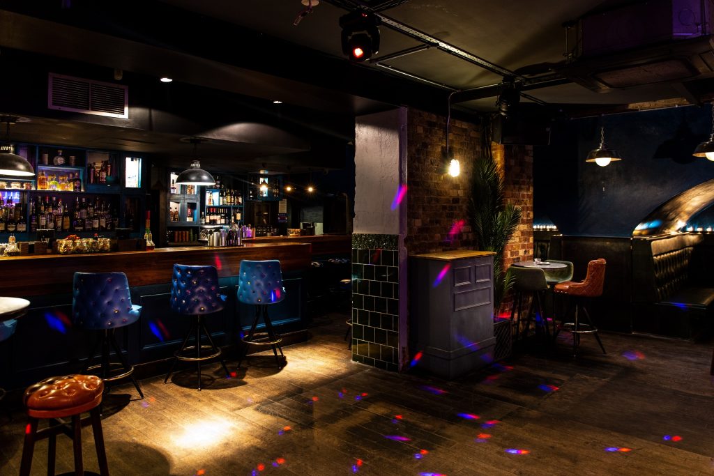 A dimly lit underground bar with eclectic blue chairs, exposed brick walls, and subtle disco lights, exuding a classic and intimate vibe.