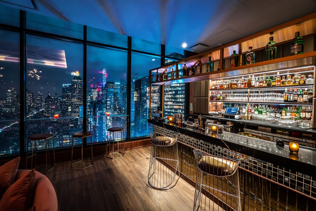 Chic rooftop bar with panoramic night views of New York City, featuring elegant seating and a well-stocked bar.
