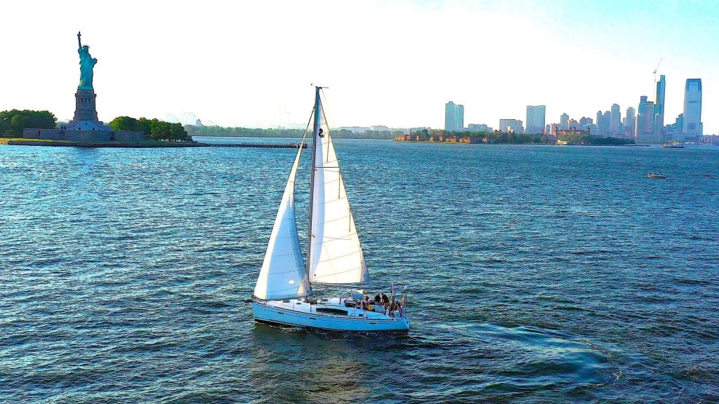 A luxury sailboat with white sails gliding on water near the Statue of Liberty with a city skyline in the background.