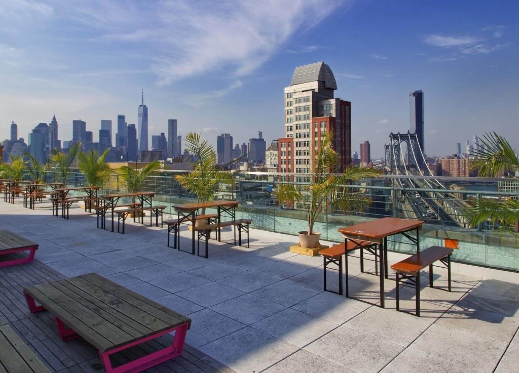 Sunny rooftop terrace with casual wooden benches and lush greenery, with a skyline backdrop including the Williamsburg Bridge.