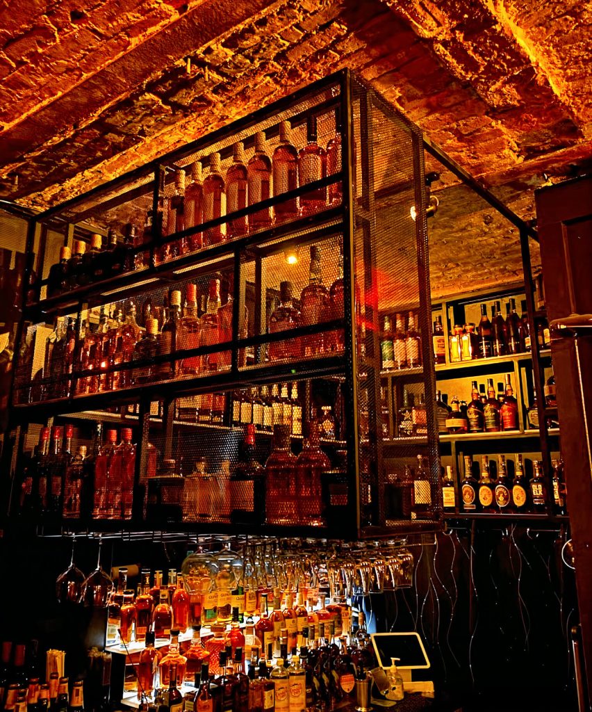 A cozy whiskey cellar with exposed brick ceiling and an extensive collection of whiskey bottles on mesh metal shelves.