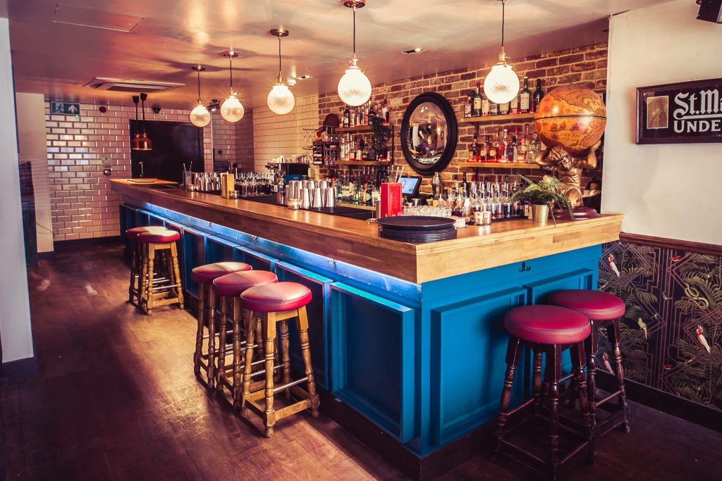 A vibrant bar with a blue and wood bar counter, tiled backsplash, and eclectic stools, creating a casual yet stylish drinking environment.