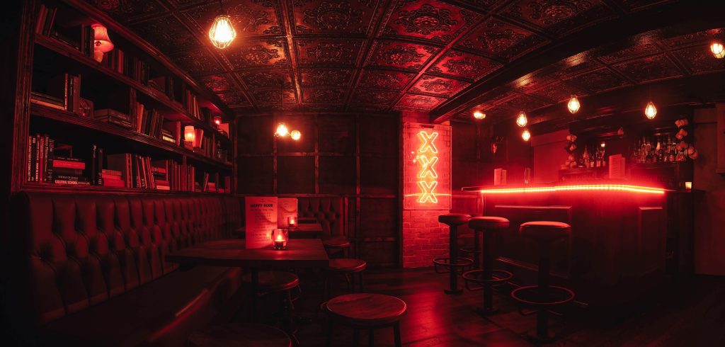A dark, moody room with red neon lighting, leather banquettes, and bookshelves filled with red books, providing an intimate speakeasy feel.