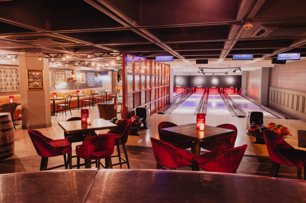 A modern bowling alley with sleek lanes, plush seating areas, and a relaxed dining space, combining entertainment and leisure in an urban setting.