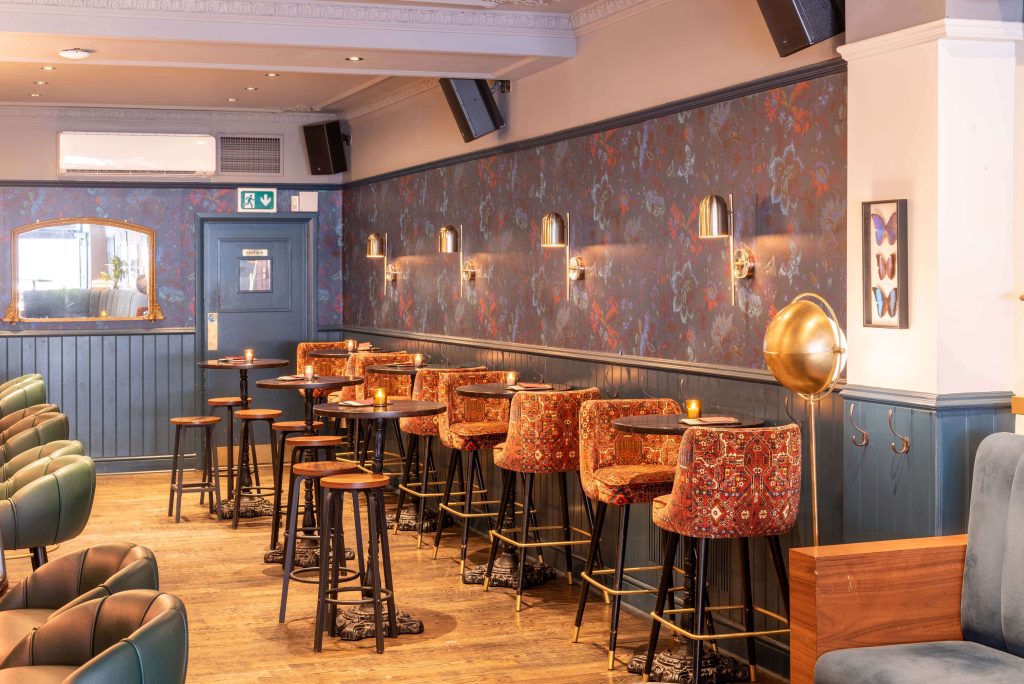 A cosy bar area with blue wainscoting, patterned wallpaper, and unique hanging lamps over patterned stools, providing a chic and inviting space.