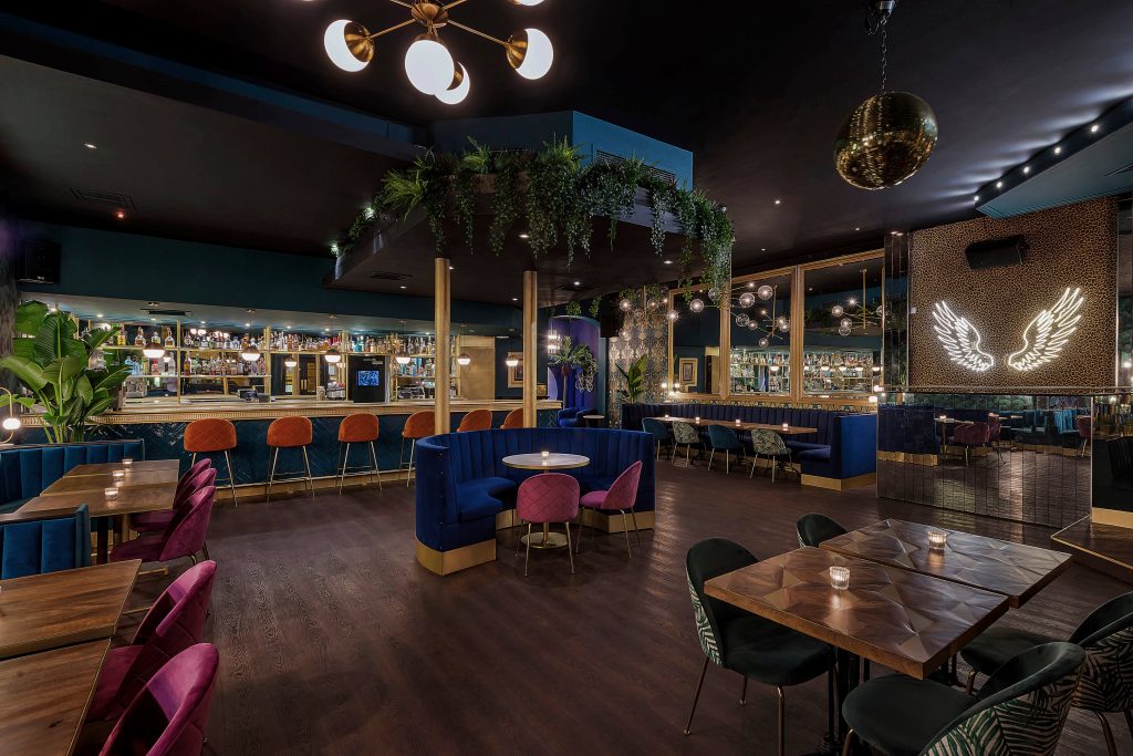 A trendy bar with dark blue walls, plush blue and pink seating, a disco ball, and a bar with a glowing winged emblem, creating a stylish, modern atmosphere.