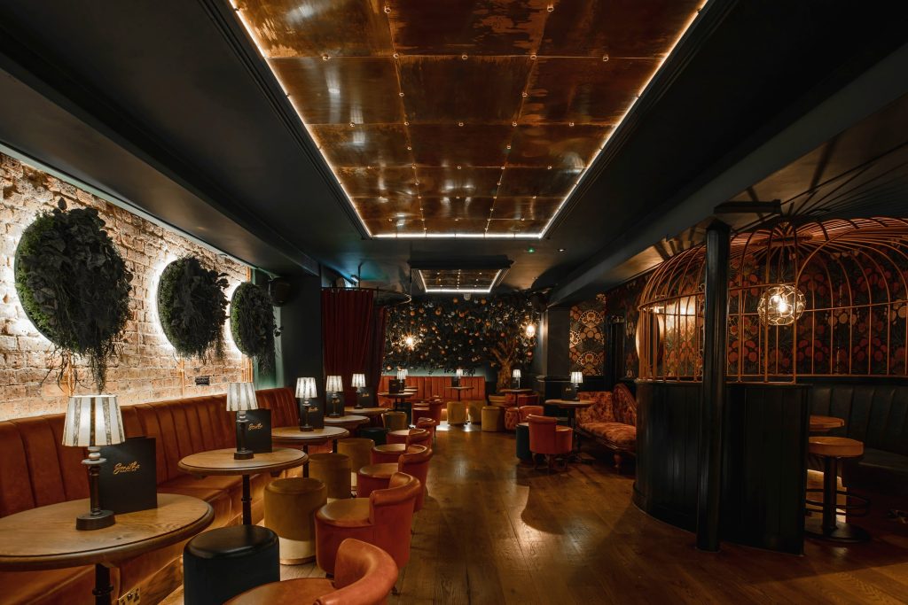 A chic cocktail bar with luxurious copper ceiling, exposed brick walls adorned with foliage, and opulent birdcage booths, creating an atmospheric setting.