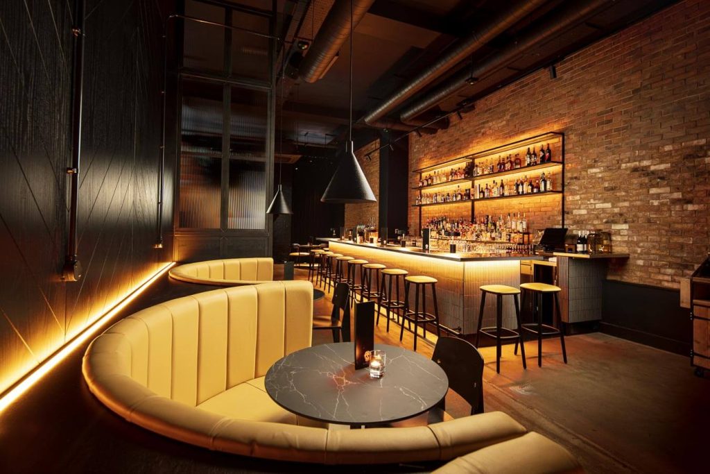 A sophisticated bar with sleek modern decor, featuring round booths, pendant lighting, and a warm wood-toned bar, creating an upscale ambience.
