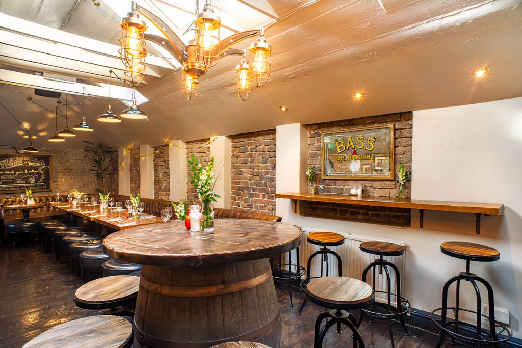 A rustic loft space with exposed beams, hanging industrial lights, and wooden barrels, offering a warm and inviting pub atmosphere.