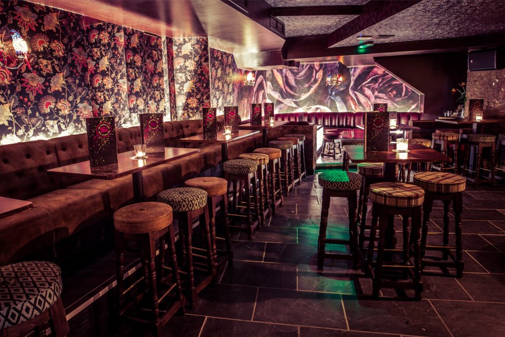 A moody bar with luxurious velvet booths, patterned wallpaper, and dim, warm lighting, providing an intimate setting for cocktails.