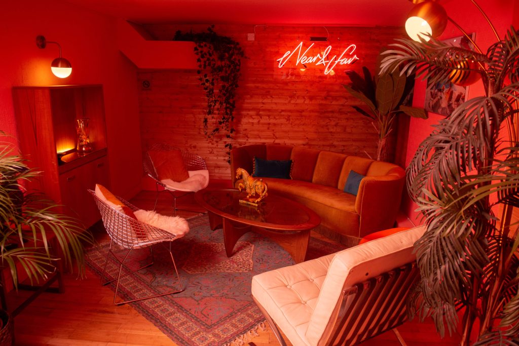 An intimate lounge with warm red lighting, plush seating, and vibrant indoor plants, offering a cosy, bohemian space for relaxation.