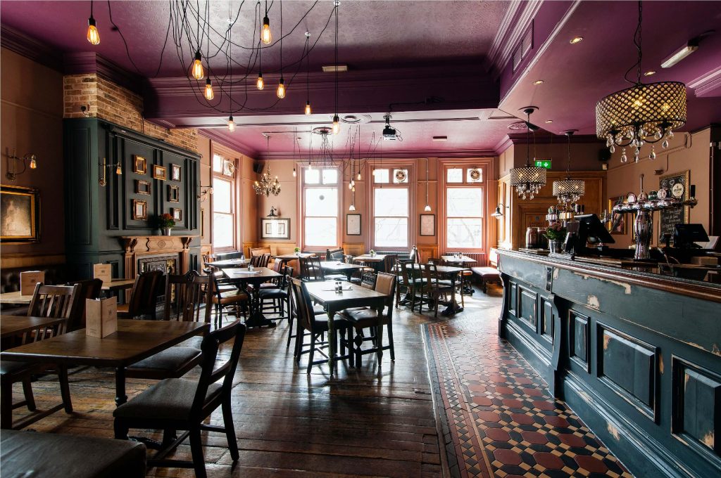 A traditional pub with purple walls, a dark wood bar and matching tables, and elegant chandeliers, offering a classic British pub experience.