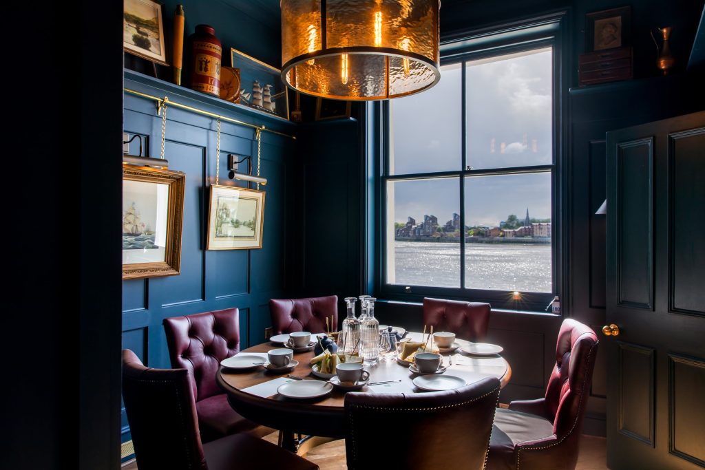 A sophisticated dining space with dark blue walls, leather chairs, and a view of the river, illuminated by a striking gold pendant light.