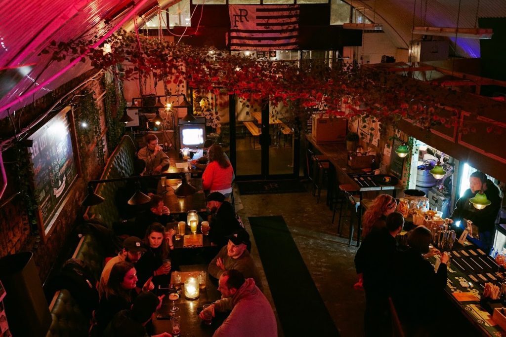 A lively taproom with an industrial ceiling, hanging greenery, and a bustling bar scene, ideal for a casual night out.