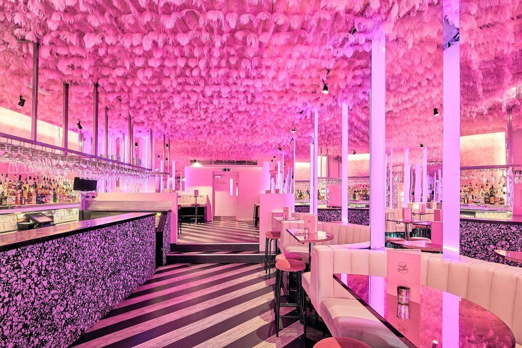 A vibrantly pink-themed bar with plush seating and a feathered ceiling, offering a trendy and lively atmosphere for socialising.