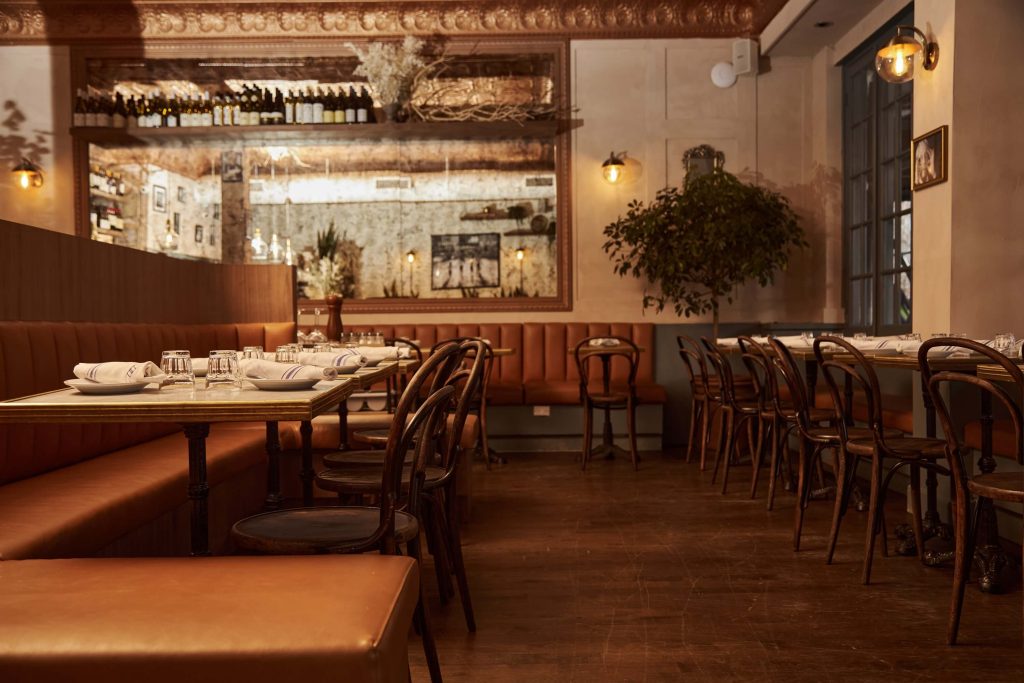 A cozy restaurant interior with a long dining table set for a meal, leather banquettes, and warm lighting reflecting off a collection of wine bottles on the wall.