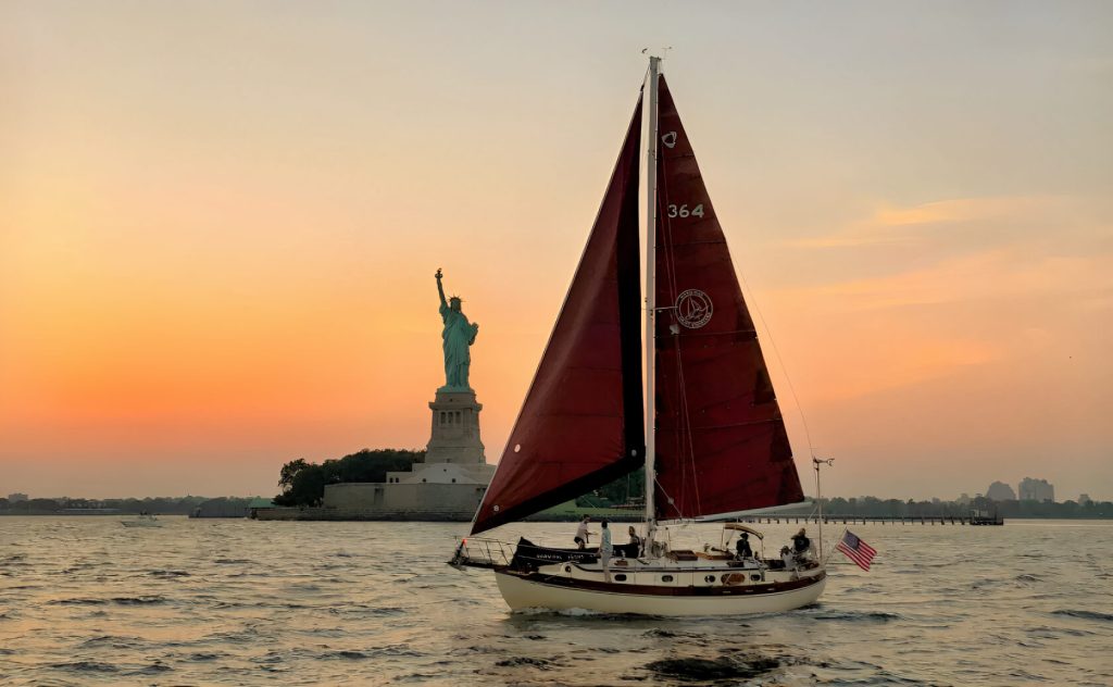 A sailboat with a red sail gliding on water near a large statue on an island at sunset, with a soft orange sky in the background.