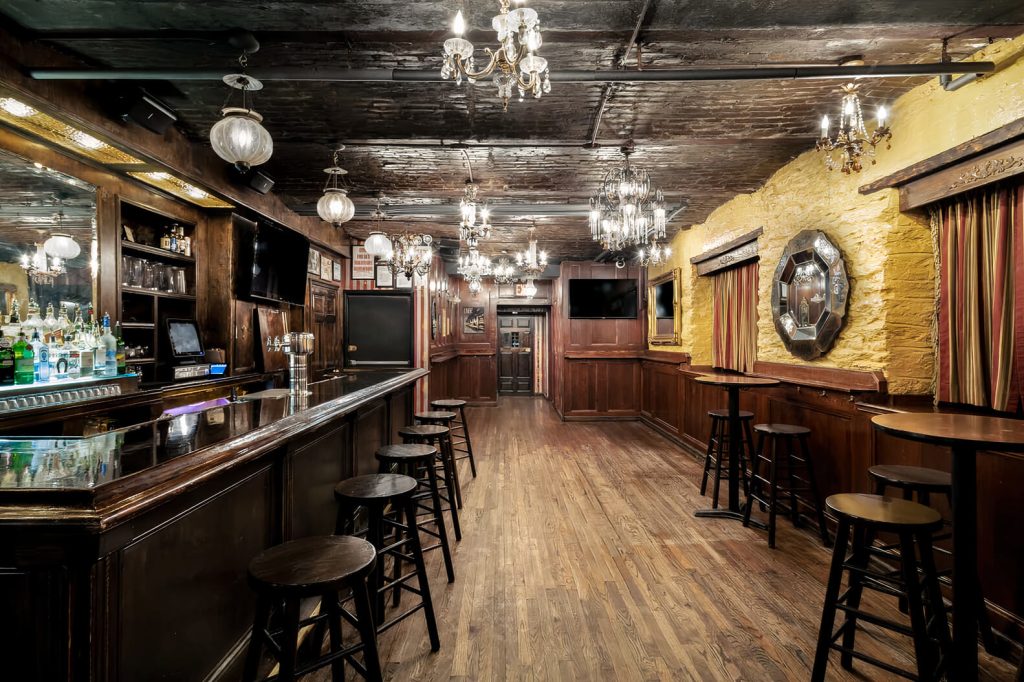 A traditional bar room with wood-paneled walls, a polished counter, vintage lighting fixtures, and several bar stools lined up along the bar.