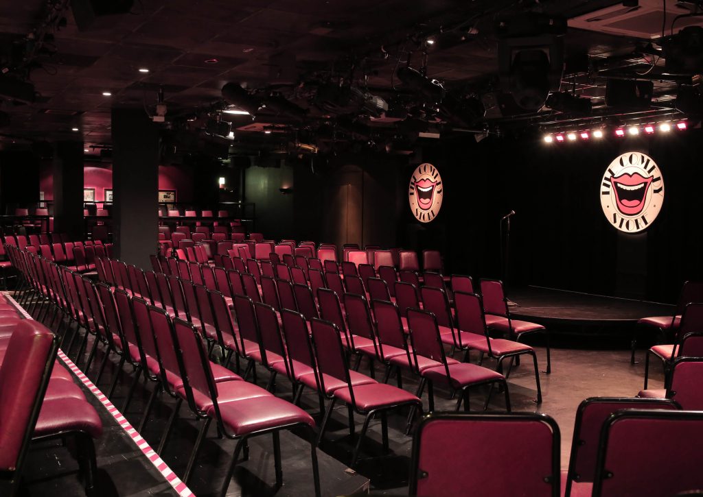 Rows of red seats facing an empty stage with comedy masks at The Comedy Store in Soho, London, set for a stag do night of laughter