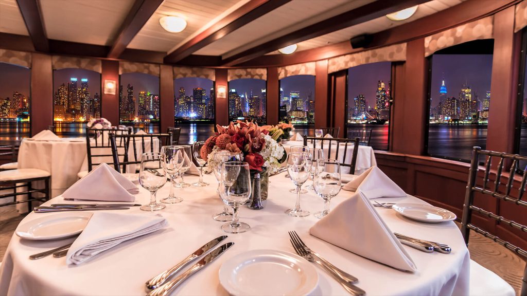 An upscale dinner party setting in NYC with tables set against large windows showcasing a nighttime view of the city skyline.