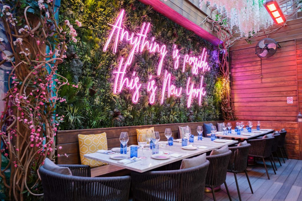 An outdoor NYC dinner party venue with vibrant neon signage, plush seating, and a lush wall garden, creating a whimsical atmosphere.