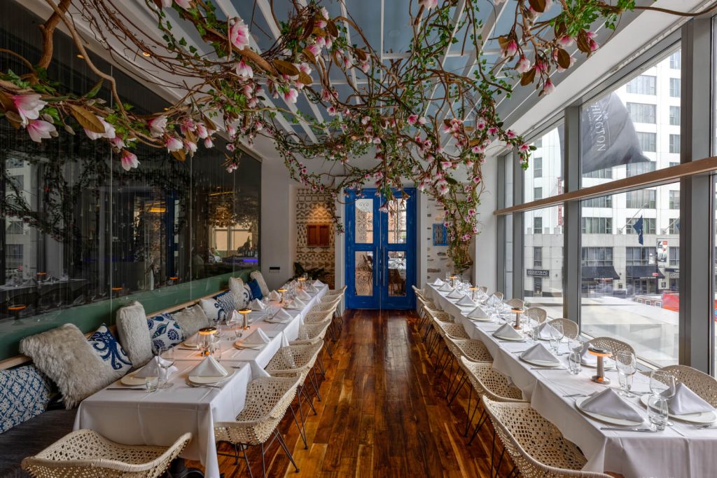 A bright and airy NYC dinner party venue with a long wooden table, white chairs, and blossoming branches overhead, invoking Greek vibes.