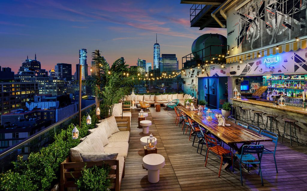 A trendy rooftop venue in NYC for a dinner party, featuring wooden benches, colorful chairs, and string lights with a cityscape in the background.