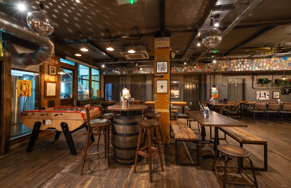 Cosy pub ambiance at Temple Brew House in London, featuring a foosball table, wooden barrel tables, and eclectic decorations for a laid-back stag party