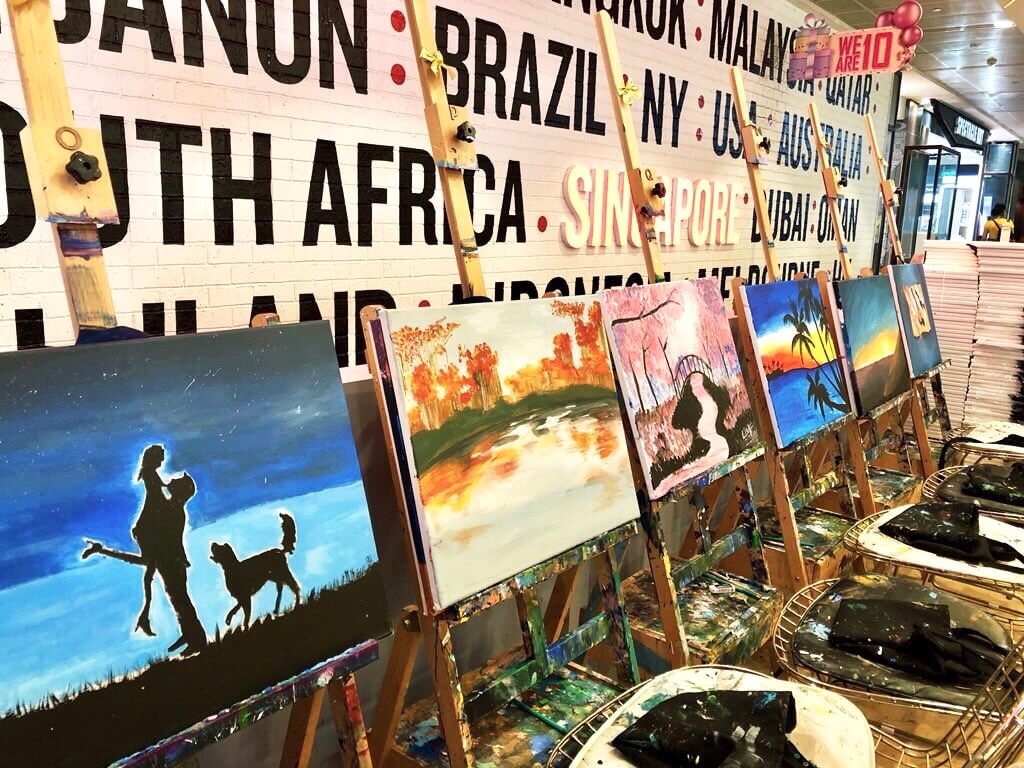 A row of easels with colourful paintings in an art studio with a wall decorated with destination names in the background.