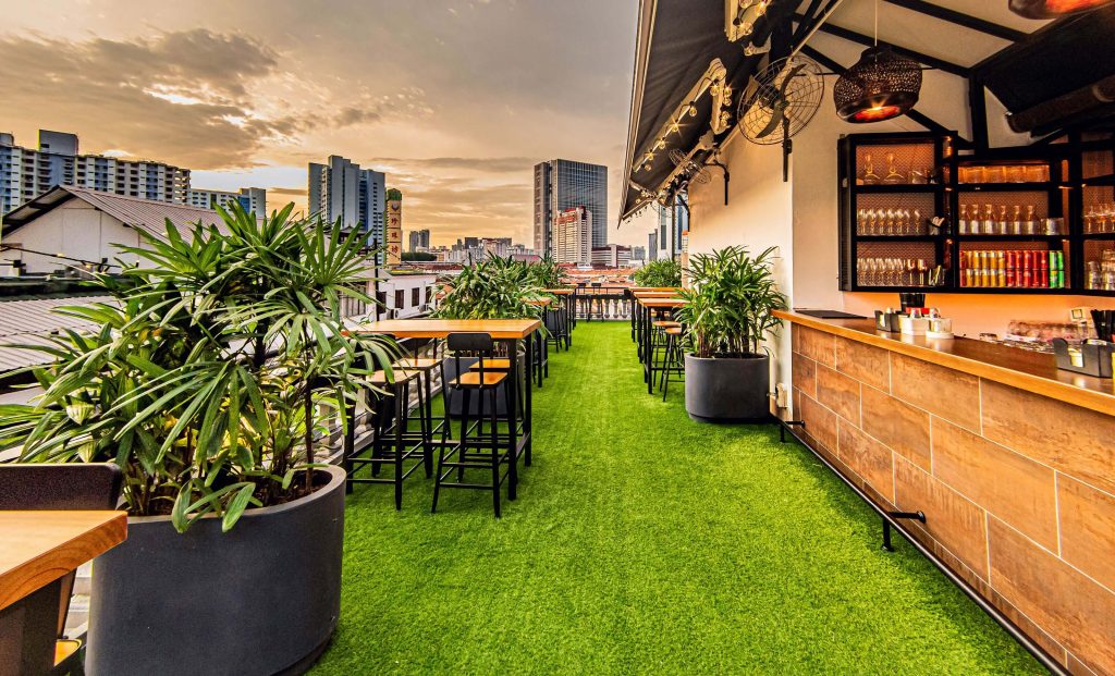 Rooftop bar setting with artificial grass floor, wooden tables, and a view of the city skyline during sunset.