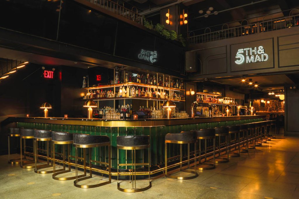 A chic bar with a dark, moody atmosphere, featuring a green marble countertop and brass detailing. The shelves are well-stocked with a variety of liquor bottles, illuminated by warm, under-shelf lighting. A neon sign that reads 