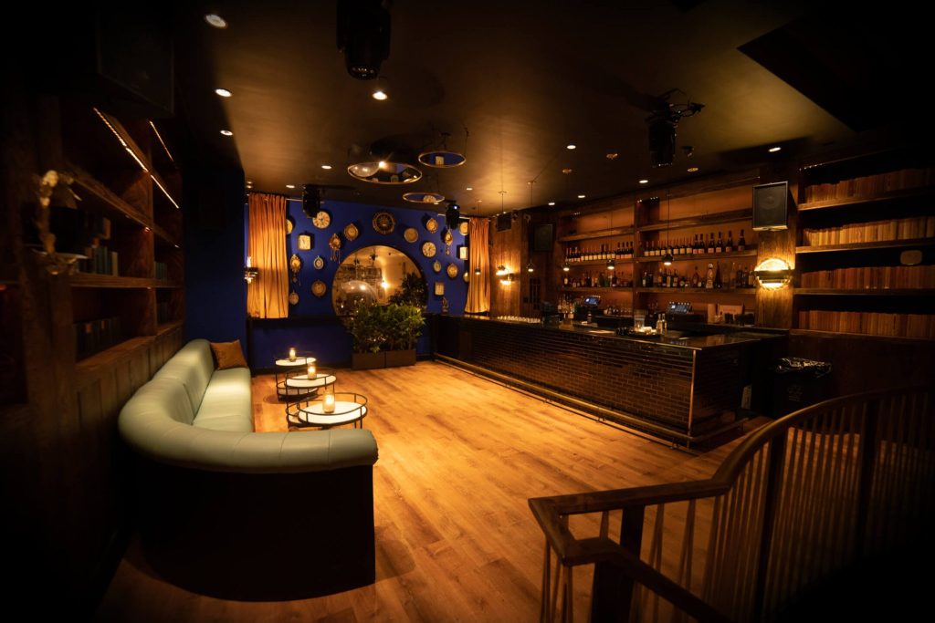 A cozy and inviting bar with a warm, dimly lit ambiance. The left side features a curved, light blue sofa with round coffee tables holding candles. A bold blue accent wall decorated with plates and mirrors contrasts with the rich wooden bookshelves to the left. The bar area on the right has a black countertop with a brick base, wooden shelves filled with bottles and books, and a glassware rack above. The space has a hardwood floor and a relaxed, sophisticated atmosphere.