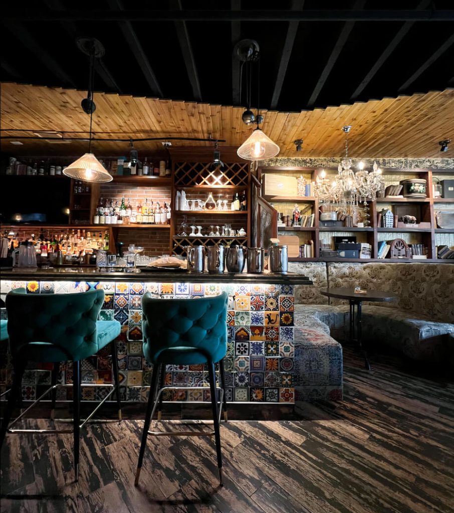 A rustic and cozy bar with eclectic tiles and plush teal bar stools. The ceiling is wooden, and the bar is adorned with hanging glass pendant lights and a chandelier. Shelves behind the bar display an assortment of bottles and decorative items.