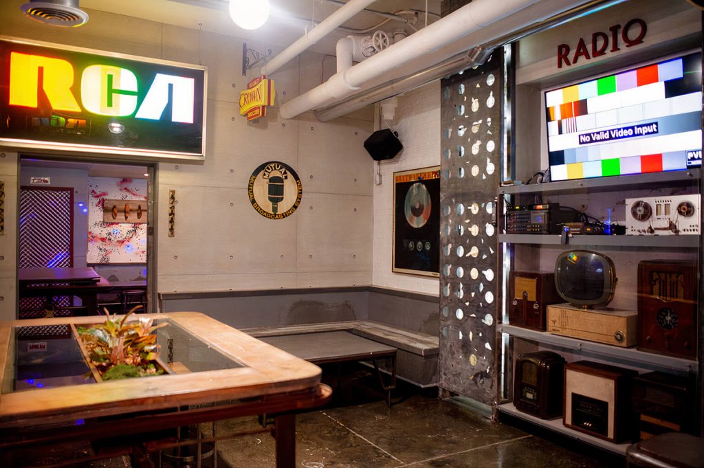 A unique, retro-styled lounge with vintage radio and television equipment. The space has a casual, industrial vibe with a white painted brick wall and a mix of bench and table seating.