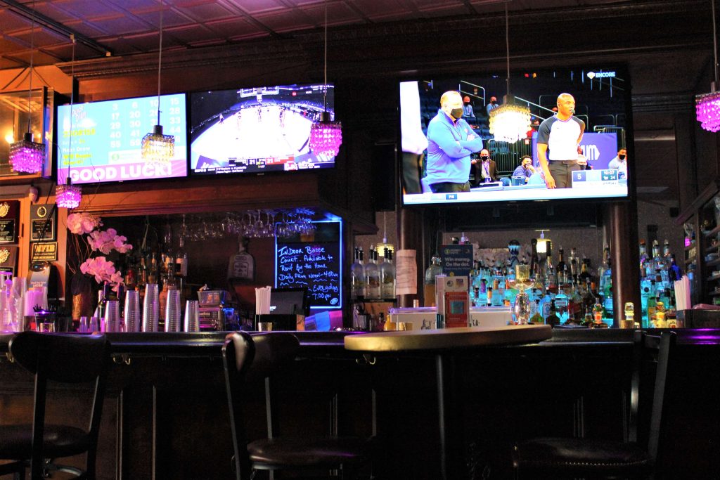 A traditional sports bar with multiple televisions displaying basketball games. The bar is lit with neon lights and adorned with sports memorabilia. Patrons can enjoy drinks at the wooden bar with black stools.