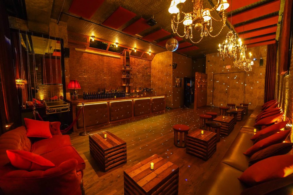 An intimate lounge with a warm, reddish ambiance and exposed brick walls. The space is decorated with chandeliers, red drapery, and cushioned benches along the walls. The wooden floor has scattered tables and the bar has a reflective surface.