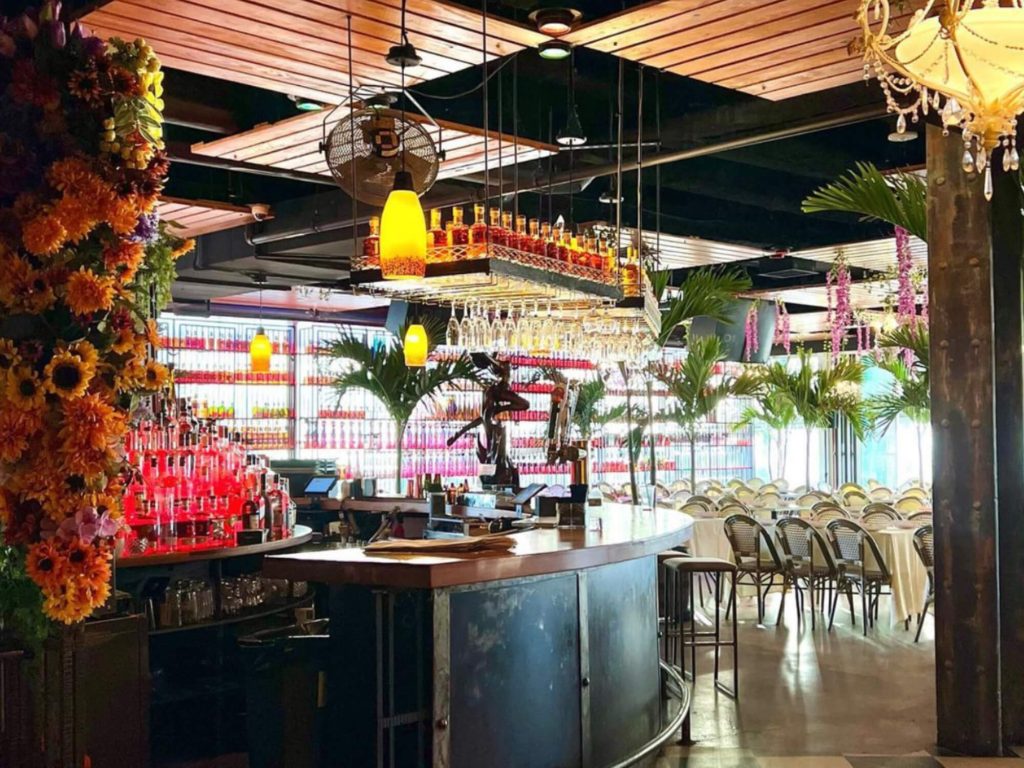 A vibrant and colorful bar with a tropical theme. The ceiling is adorned with lush floral arrangements and pendant lights. The back wall is stacked with bright red bottles, and the space has a casual, airy feel with plenty of seating.