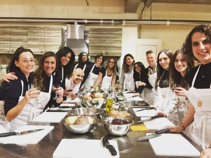 A group of smiling people wearing aprons and holding wine glasses during a cooking class, a popular group activity in NYC.
