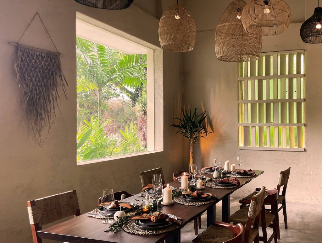 An intimate dining area with tables set for a meal, rustic chairs, hanging wicker lampshades, and a view of lush greenery outside a large window.